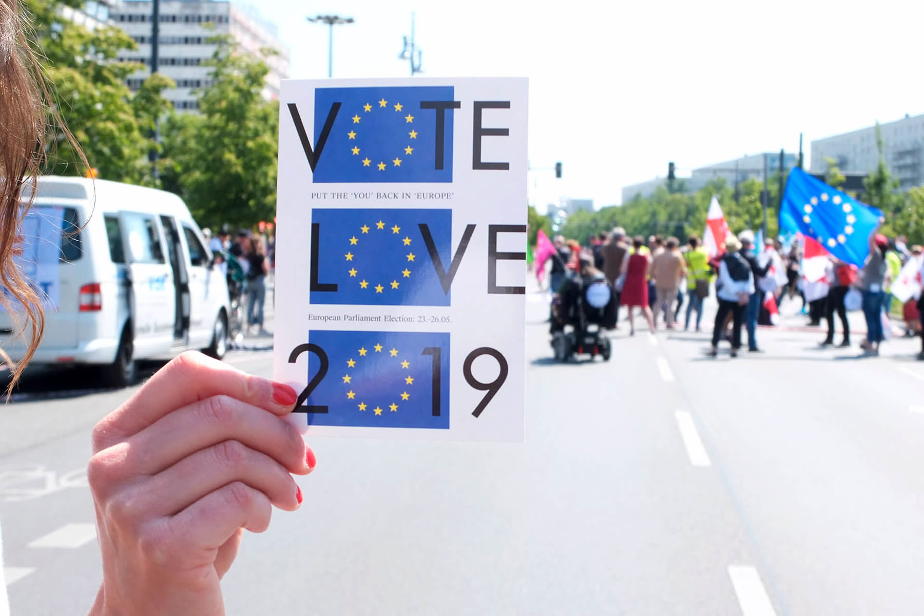 Hand holding a Vote Love 2019 card in front of a march with European flags